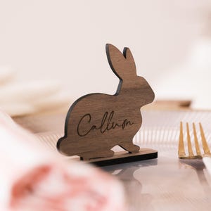 May include: Wooden bunny rabbit place card with the name "Callan" engraved on it. The place card is sitting on a table with a white tablecloth and a gold fork.