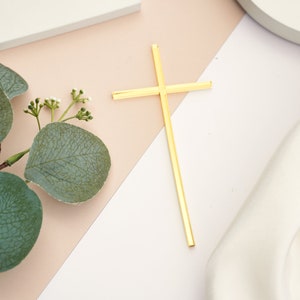 May include: A gold cross made of metal, on a white and beige background. The cross is simple and elegant, with a smooth finish.
