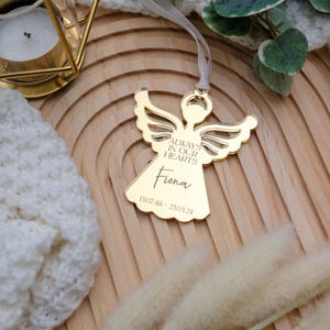 May include: A gold angel-shaped ornament with the inscription "ALWAYS IN OUR HEARTS" and the name "Fiona". The dates 13.07.48 - 23.03.24 are also engraved. The ornament is suspended by a white ribbon.