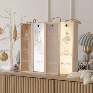 Può includere: Quattro scatole di vino in legno con manici in corda, ognuna con un design e un nome diversi: Jessica, Nancy, Gemma e Harvey. Le scatole hanno un tema natalizio con disegni di albero, renna e fiocco di neve. Sono visibili anche le parole "Merry Christmas".
