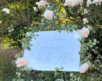 Puede incluir: Un cartel de bienvenida azul claro con letras blancas. El cartel dice "Bienvenido a nuestra boda Cadi & Alex 5 de agosto de 20". El cartel está decorado con flores blancas y vegetación.