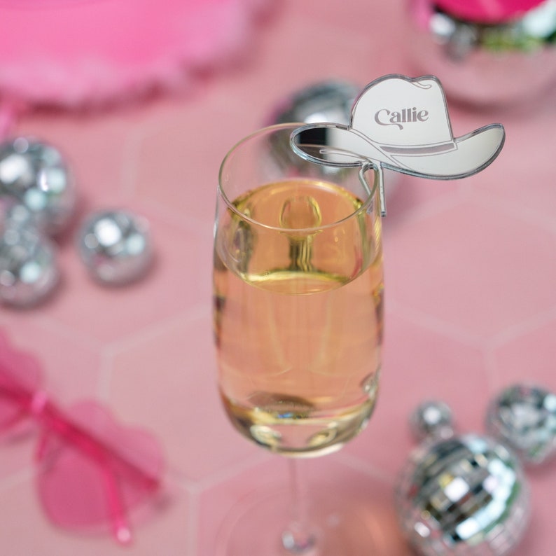 May include: A silver mirrored cowboy hat with the name "Callie" engraved on it. The hat is placed on the rim of a champagne flute.