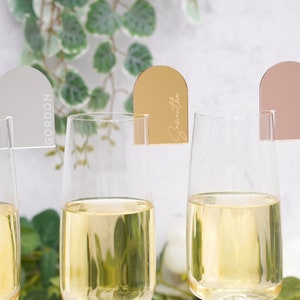 May include: Three champagne flutes with personalized acrylic place card holders. The place cards are in the shape of an arch and are in silver, gold, and rose gold. The names "Gordon", "Samantha", and "Hannah" are written on the place cards.