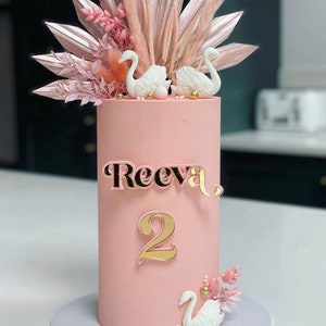 Könnte beinhalten: Eine rosafarbene zweistöckige Torte, verziert mit weißen Schwänen, getrockneten Blumen und goldenen Akzenten. Die Torte ist mit einer goldenen Zahl "2" und dem Namen "Reeva" in goldenen Buchstaben verziert.