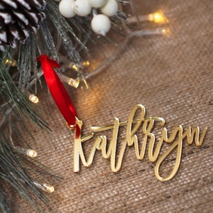 Puede incluir: Adorno navideño de acrílico dorado con el nombre "Kathryn" escrito en cursiva. El adorno está colgado de una cinta roja.