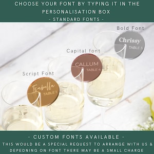 May include: Three clear glass champagne flutes with gold, silver and brown acrylic table number place cards. The place cards have different fonts and the text "Chrissy Table 3", "Callum Table 4", and "Isabella Table 1".