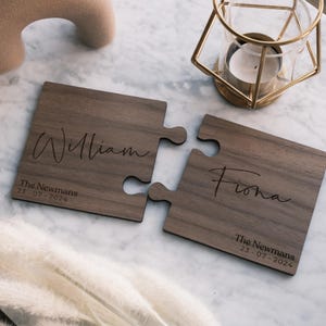 Puede incluir: Dos posavasos de madera con forma de piezas de rompecabezas. Uno está grabado con el nombre "William" y el otro con "Fiona". Ambos incluyen "The Newmans" y la fecha "23-07-2024". Los posavasos son de color marrón oscuro.