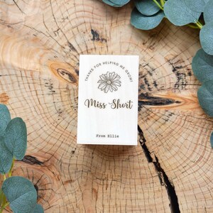 May include: A small wooden sign with the text "Thanks for helping me grow! Miss Short From Ellie" and a simple flower illustration.