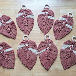 May include: Eight handmade macrame coasters in a leaf design. The coasters are a dusty rose color with a white accent stripe. Each coaster has a wooden ring attached for hanging or carrying. The coasters are arranged on a wooden surface.