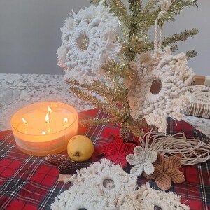 May include: A festive display featuring handmade macrame snowflake ornaments in various sizes and colors, including white, red, and brown. A lit candle, a pear, and decorative corn are arranged on a red plaid surface, with a small gold tinsel tree in the background.