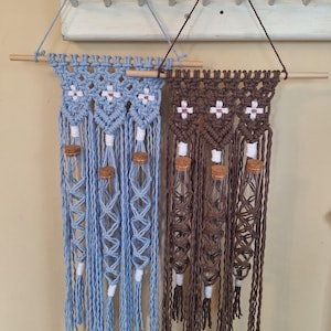 May include: Two macrame wall hangings with wooden dowels and glass test tubes. The left hanging is light blue with white tassels and the right hanging is brown with white tassels.