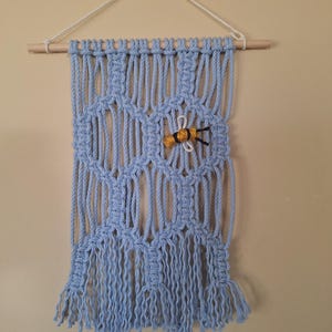 May include: A light blue macrame wall hanging with a wooden dowel and a white string for hanging. The macrame features a geometric design and a small, yellow and black bee accent. The bottom of the hanging has a fringe.