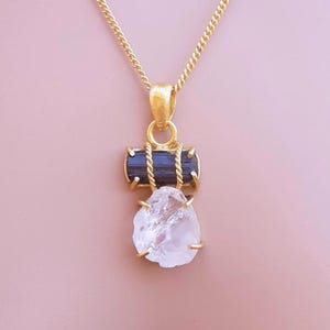 May include: A gold chain necklace with a pendant. The pendant has a dark blue-black rectangular stone above a clear, irregular-shaped stone, both set in gold-coloured metal. The necklace is displayed against a pink background.