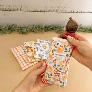 May include: Several fabric eyeglass cases with different patterns. One case holds sunglasses. The cases feature floral, teddy bear, and plaid designs. The case in focus has a floral pattern with the text "Cotton Daisies".