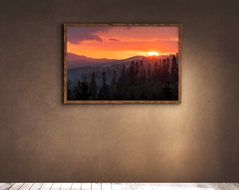 Könnte beinhalten: Ein gerahmtes Foto eines Sonnenuntergangs über einem Wald mit einem Gebirge im Hintergrund. Der Himmel ist leuchtend orange und rosa, und die Sonne geht hinter den Bäumen unter.