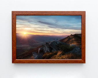 Könnte beinhalten: Ein gerahmtes Foto einer Bergkette bei Sonnenuntergang. Die Sonne geht hinter den Bergen unter und wirft einen warmen Schein auf die Landschaft. Der Himmel ist eine Mischung aus Blau, Rosa und Orange. Die Berge sind felsig und zerklüftet, mit einigen Bäumen, die an den Hängen wachsen.