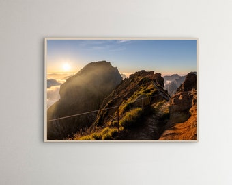 Könnte beinhalten: Gerahmtes Landschaftsfoto eines Bergpanoramas bei Sonnenaufgang. Die Sonne scheint hell und wirft einen goldenen Schein auf die Gipfel und einen Weg mit Seilgeländer. Der Himmel wechselt von Blau zu Orange.
