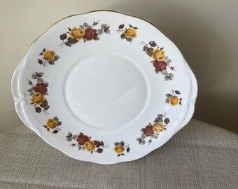 May include: A white cake plate with a gold rim and a floral design featuring yellow and brown roses. The plate has a scalloped edge.