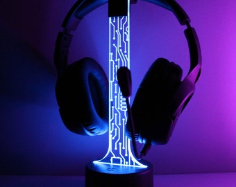 May include: A black headset sits on a blue illuminated headphone stand. The stand has a circuit board design and a round base. The background is a gradient of purple and blue.