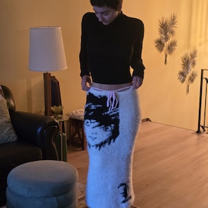 Könnte beinhalten: Ein langer, flauschiger weißer Rock mit einem markanten schwarzen Grafikdruck eines Gesichts auf der Vorderseite. Der Rock hat ein hoch tailliertes Design mit einem dünnen, hellrosa Kordelzug in der Taille, der einen gerafften Effekt erzeugt.