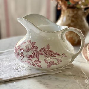 May include: A cream-colored ceramic pitcher featuring a wide spout and a curved handle. The exterior is decorated with a detailed maroon-colored transferware pattern depicting a small bird perched on a branch surrounded by blooming flowers.