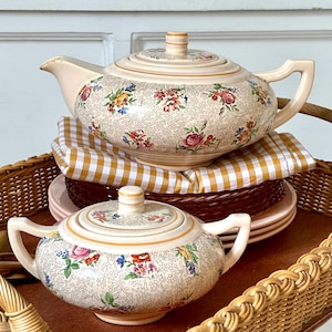 Peut inclure: Une théière en céramique et un sucrier assorti avec couvercle, présentant une base couleur crème ornée d'un motif floral brun dense et complexe avec des accents de roses colorées. Les deux pièces ont une forme arrondie et des anses.