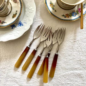 Puede incluir: Un conjunto de seis tenedores antiguos con púas de acero inoxidable y mangos que transicionan de marrón oscuro a ámbar claro. Los tenedores están dispuestos sobre un mantel blanco, con porcelana con motivos florales en el fondo.
