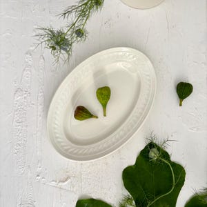 Könnte beinhalten: Eine ovale Servierplatte in Weiß mit dekorativem Rand, die zwei grüne Feigen und eine teilweise braune Feige enthält. Grüne Blätter und Zweige mit Grünzeug sind um die Platte herum auf einer strukturierten weißen Oberfläche verstreut.