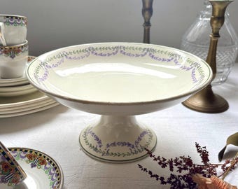 Puede incluir: Un soporte de pastel de pedestal blanco con un diseño floral verde y morado. El soporte está sobre un mantel blanco.