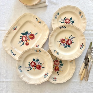 Peut inclure: Ensemble d'assiettes et un plat de service anciens de couleur crème, aux bords festonnés. Chaque pièce présente un motif floral peint à la main avec des fleurs rouges, bleues et jaunes. Des couverts avec des manches en os sont posés sur le côté.