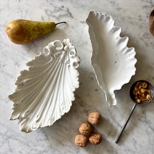 Peut inclure: Deux plats de service en céramique blancs en forme de feuille avec des détails ornés. Une poire mûre, des noix et une petite cuillère avec des cacahuètes sont disposées sur une surface en marbre, suggérant un thème décoratif ou culinaire.