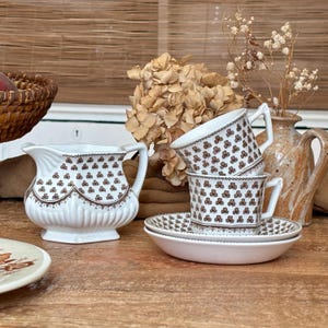 Peut inclure: Un pot à lait en céramique blanche et deux tasses à thé assorties avec soucoupes, ornés d'un motif répétitif de trèfles bruns. Le pot à lait présente un bord festonné et une base côtelée, tandis que les tasses ont des anses angulaires.