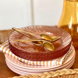 Peut inclure: Une pile d'assiettes en céramique rose clair surmontée d'une pile d'assiettes en verre ambré translucide avec un bord texturé. Deux cuillères en métal doré reposent sur l'assiette en verre supérieure, avec une serviette à carreaux vichy.