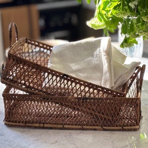 Puede incluir: Dos cestas rectangulares de mimbre marrón tejido con asas, una apilada encima de la otra. Un paño de lino blanco doblado está dentro de la cesta superior. Las cestas están sobre una superficie de mármol.