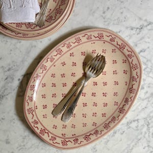 Peut inclure: Un plat de service ovale en céramique avec une base de couleur crème, orné d'un motif répétitif de petites fleurs rouges et d'une bordure décorative rouge. Une fourchette de service en métal argenté repose au centre du plat.