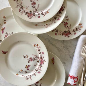 May include: A set of white ceramic plates adorned with floral and butterfly patterns. The plates showcase pink and purple flowers with brown stems and leaves. Some plates feature a butterfly motif. A white napkin with red embroidery and cutlery are also present.