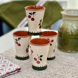 Peut inclure: Quatre gobelets en céramique avec un extérieur crème et un intérieur brun rougeâtre. Chaque gobelet est décoré d'un motif de branche de cerisier rouge et vert. La base de chaque gobelet est verte avec de petits points blancs.