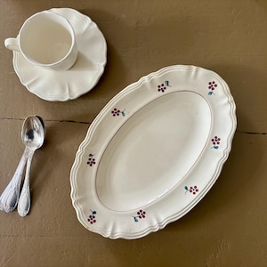 May include: Cream-colored ceramic serving platter, teacup, saucer, and spoons. The oval platter has a scalloped edge and is decorated with small floral motifs in red, blue, and green. The teacup and saucer also have scalloped edges.