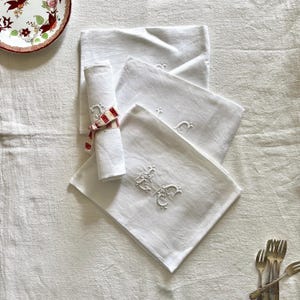 May include: A set of white linen napkins featuring intricate tone-on-tone embroidered monograms. One napkin is neatly rolled and secured with a red and white patterned ribbon. The fabric displays a visible weave texture and clean, folded edges.
