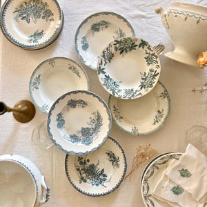Puede incluir: Una colección de platos, cuencos y fuentes antiguos blancos y azules con motivos florales. Los platos tienen un delicado diseño floral azul alrededor de los bordes y en el centro. También se ven un candelabro de latón y una fuente.