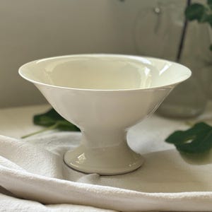 May include: A cream-coloured ceramic pedestal bowl with a wide, flared rim and a smooth, glossy finish. The bowl features a deep, rounded basin that tapers down to a sturdy, circular base, creating a classic silhouette for serving or display.