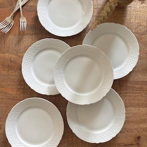 Puede incluir: Un juego de cinco platos de cerámica blanca con un delicado patrón floral y de celosía en relieve a lo largo de los bordes festoneados, acompañado de dos tenedores de metal plateado con mangos ornamentados, dispuestos sobre una superficie de madera.
