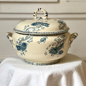 Peut inclure: Soupière en céramique crème avec couvercle, ornée de motifs floraux bleus. Le plat est de forme ronde, avec deux poignées latérales et une anse sur le couvercle. Le bord et la base présentent une bordure pointillée.
