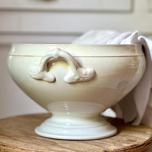 Puede incluir: Cuenco de cerámica color crema, de estilo antiguo, con dos asas ornamentadas. El cuenco tiene una forma ancha y redondeada y se apoya sobre una base de pedestal. Un paño blanco es parcialmente visible en el interior. Para servir.