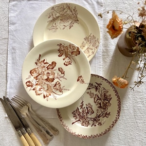 Peut inclure: Trois assiettes anciennes en céramique avec des motifs floraux bruns et crème. Les assiettes sont disposées sur une nappe blanche, accompagnées d'argenterie vintage. Chaque assiette présente un motif floral unique, idéal pour une table vintage.