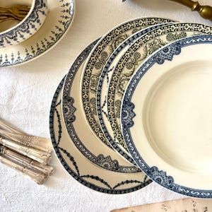 Könnte beinhalten: Eine Sammlung antiker cremefarbener Teller mit aufwendigen blauen Blumen- und geometrischen Mustern. Die Teller sind auf einer weißen Tischdecke angeordnet, zusammen mit einer passenden Schüssel und Silberbesteck, was eine Vintage-Ästhetik erzeugt.