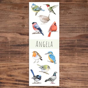 Puede incluir: Un marcapáginas blanco con ilustraciones de acuarela de varias aves, incluyendo un herrerillo común, un colibrí y un cardenal. El nombre "ANGELA" está impreso en letras mayúsculas negras. El fondo es una superficie de madera.