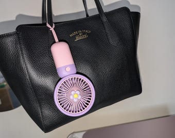 May include: A black leather tote bag with the text "MADE IN ITALY BY Gucci" in gold lettering. A small, handheld fan in pink and purple hangs from the bag. The fan has a flower-shaped center.