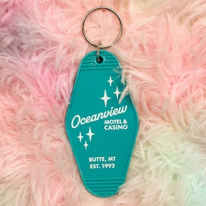 May include: Teal blue motel key chain with white text that reads "Oceanview Motel & Casino Butte, MT Est. 1992". The keychain has a retro design with five white stars.