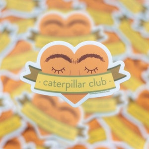 May include: A sticker with a heart-shaped design featuring a closed-eye face with eyebrows. The heart is orange and the eyebrows are brown. A yellow banner with the text "caterpillar club." is wrapped around the bottom of the heart. The text "rosewatercreative.com" is printed in small font at the bottom of the sticker.